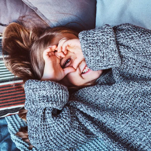 Young girl holding up her fingers in a heart shape over her eyes on a bed.