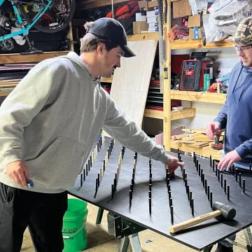 two men in a garage building hammering in short spokes to make to make a giant Plinko board 