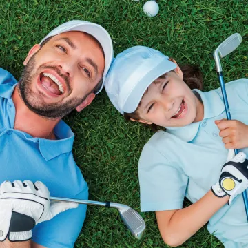 father and son laying down on grass smiling holding golf clubs