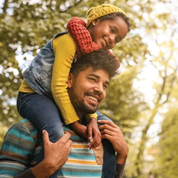 A girl riding on her dads shoulders outside with sun shining